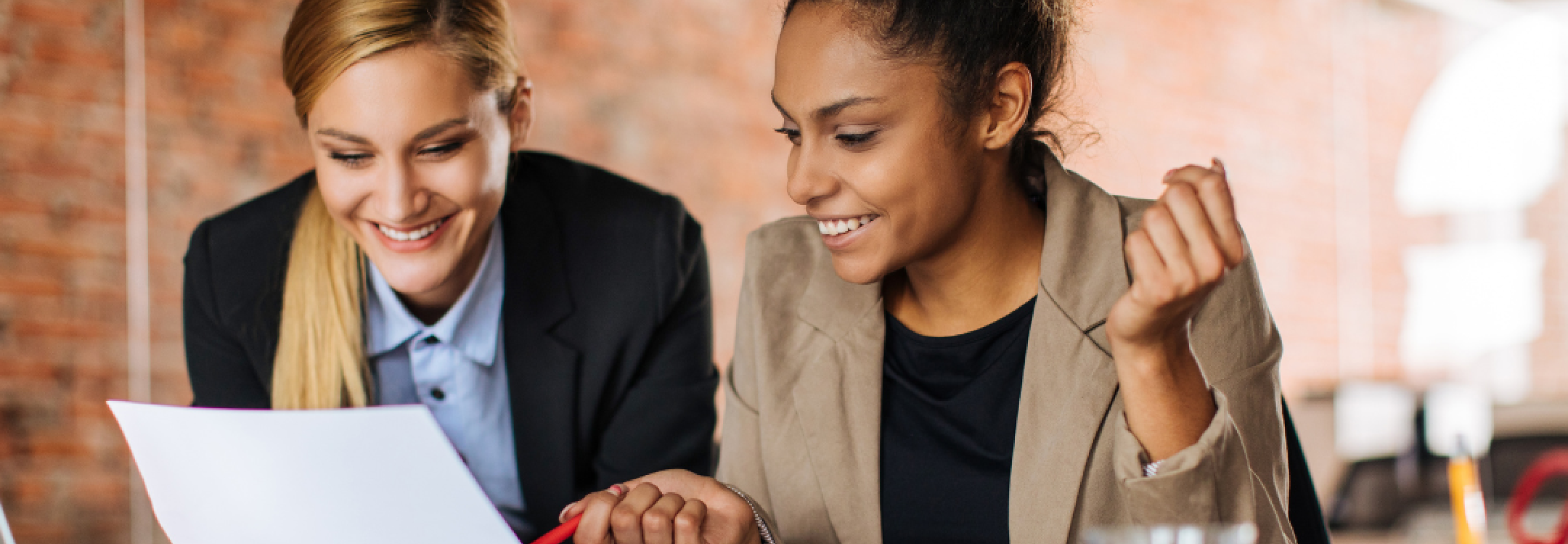 Two women in professional wear look at a laptop together and smile.