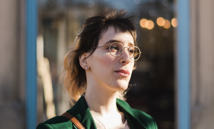 A woman in a green coat and wire framed glasses looks to her left meaningfully