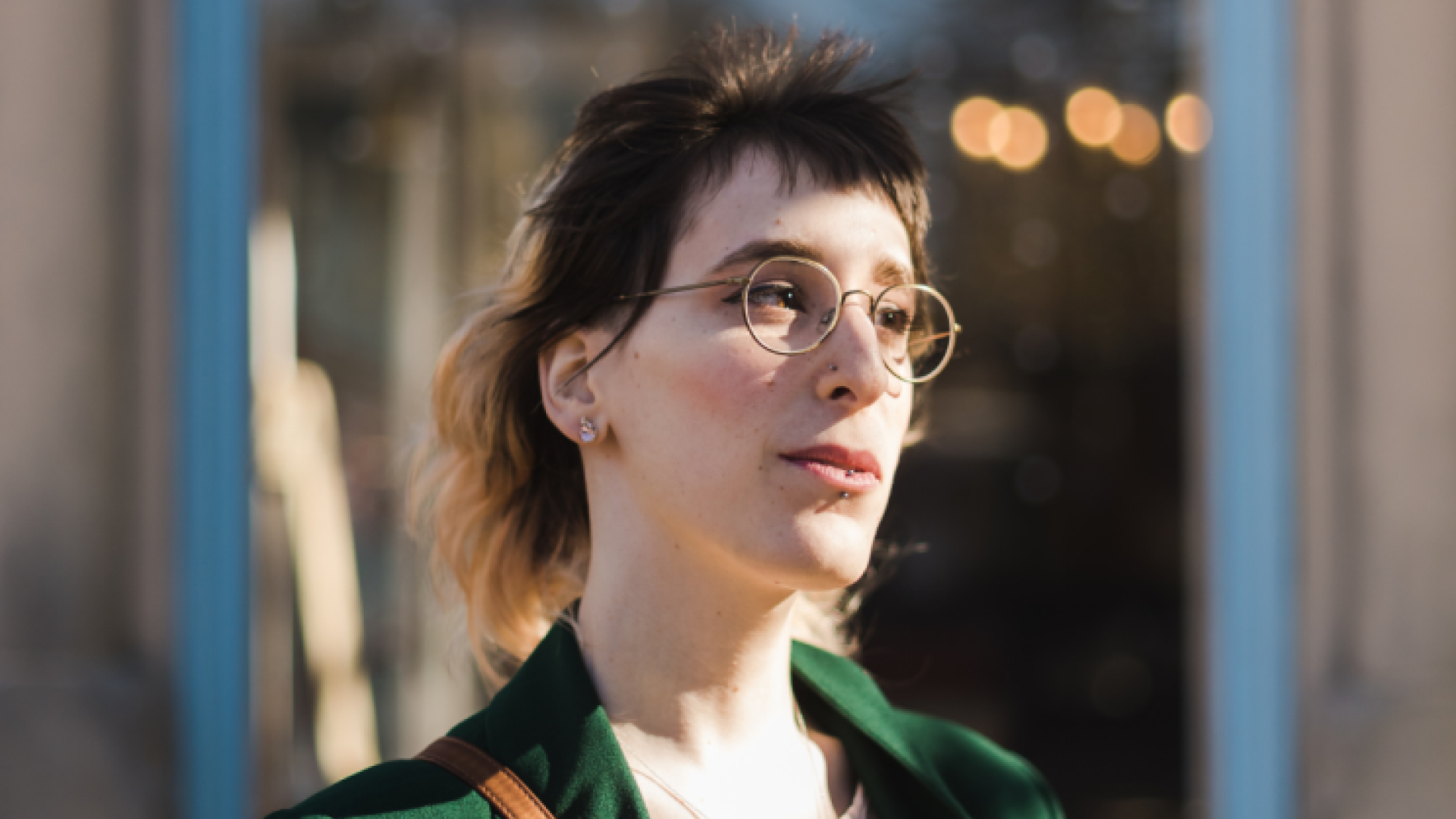 A woman with soft brown hair and glasses in a green coat poses in the foreground of the photo