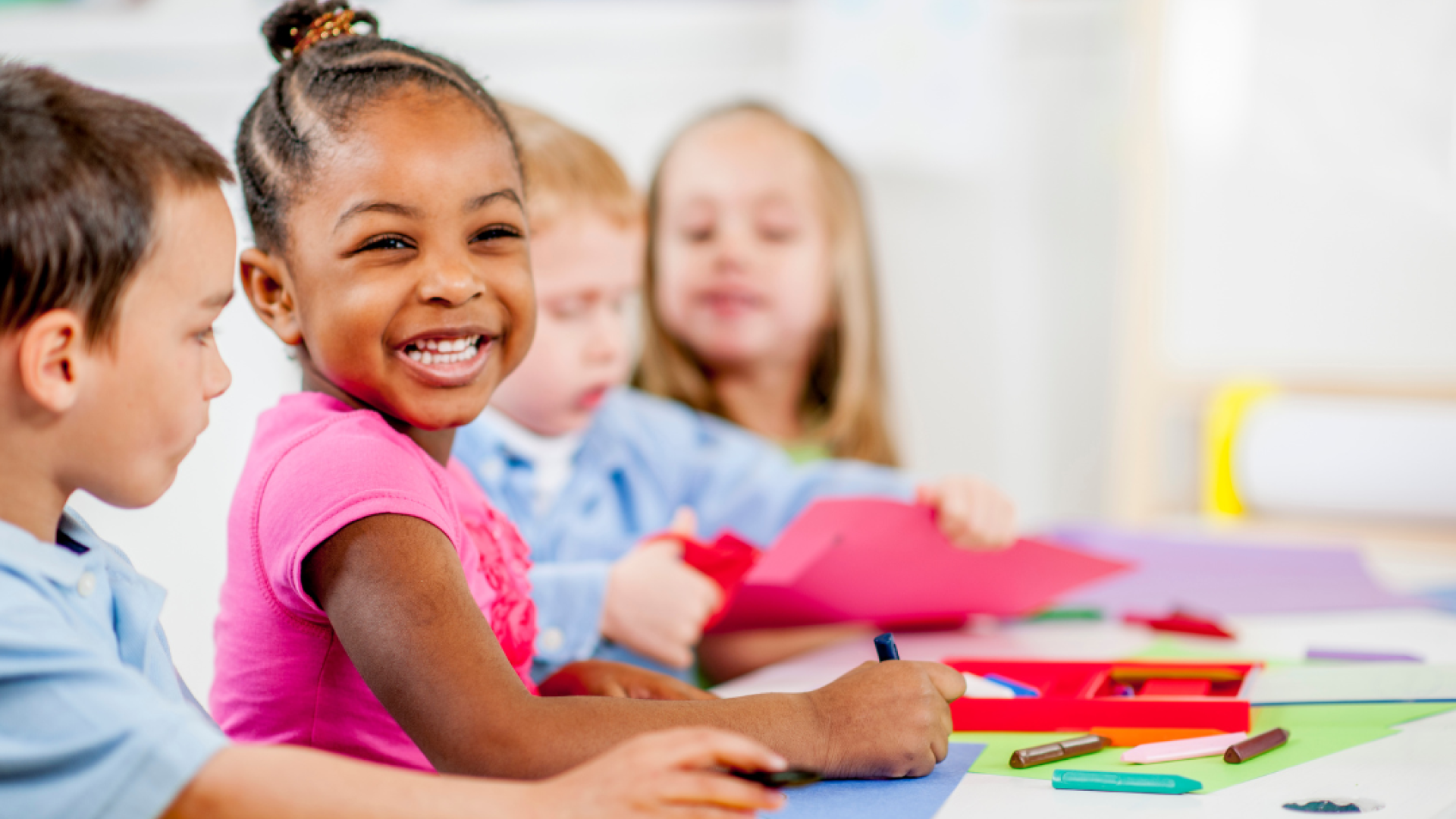 A child makes a huge grin at the camera while she colors with three other children.