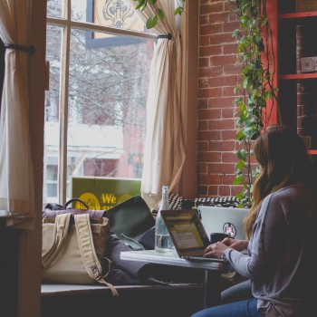 Photo of woman typing on a computer. She sits at a desk in front of a bright window in a cozy room.Photo by Bonnie Kittle on Unsplash