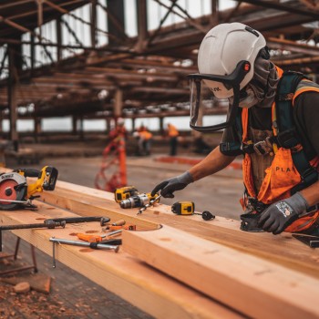 Construction worker with a hard hat and face shield on, measuring wood planks.